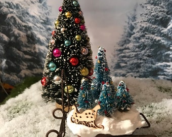 Puede incluir: Una escena navideña en miniatura con un pequeño árbol de Navidad decorado, un trineo y varios pinos más pequeños cubiertos de nieve. El trineo es de metal y tiene un recorte de madera de un trineo con la palabra "Merry" en él.