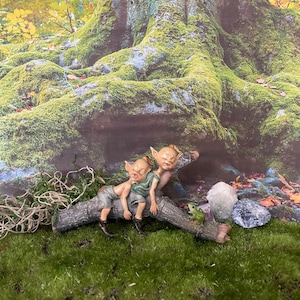 May include: Two small, green-skinned figures with pointed ears are sleeping on a brown log. A small green frog sits on the log near the figures. The log is resting on a bed of green moss.