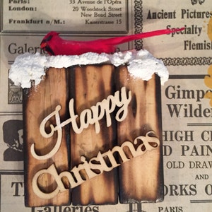 May include: A wooden sign with the words "Happy Christmas" in a rustic style. The sign is decorated with a red cardinal figurine and a snowy white top.