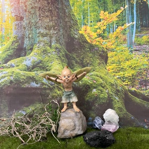 May include: A small, green-winged fairy figurine with a mischievous grin stands on a rock in a mossy woodland setting.  The fairy is surrounded by various crystals, including a large clear quartz crystal, a dark purple amethyst, and a black obsidian.