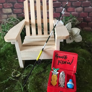 Puede incluir: Una silla de madera en miniatura con una caña de pescar apoyada en ella. Una caja de aparejos roja con las palabras "Gone Fishin'" está abierta y contiene tres peces de madera y una botella azul.