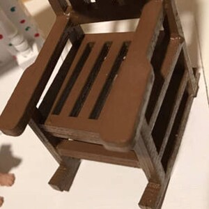 Op de afbeelding: Een bruine houten schommelstoel met een latten zitting en rugleuning. De stoel is gemaakt van onbehandeld hout en heeft een rustieke uitstraling.
