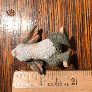 May include: A small, gray and white rabbit figurine with a pink nose. The figurine is about 1 inch tall and is lying on its back with its legs in the air. The figurine is on a wooden surface with a ruler next to it.
