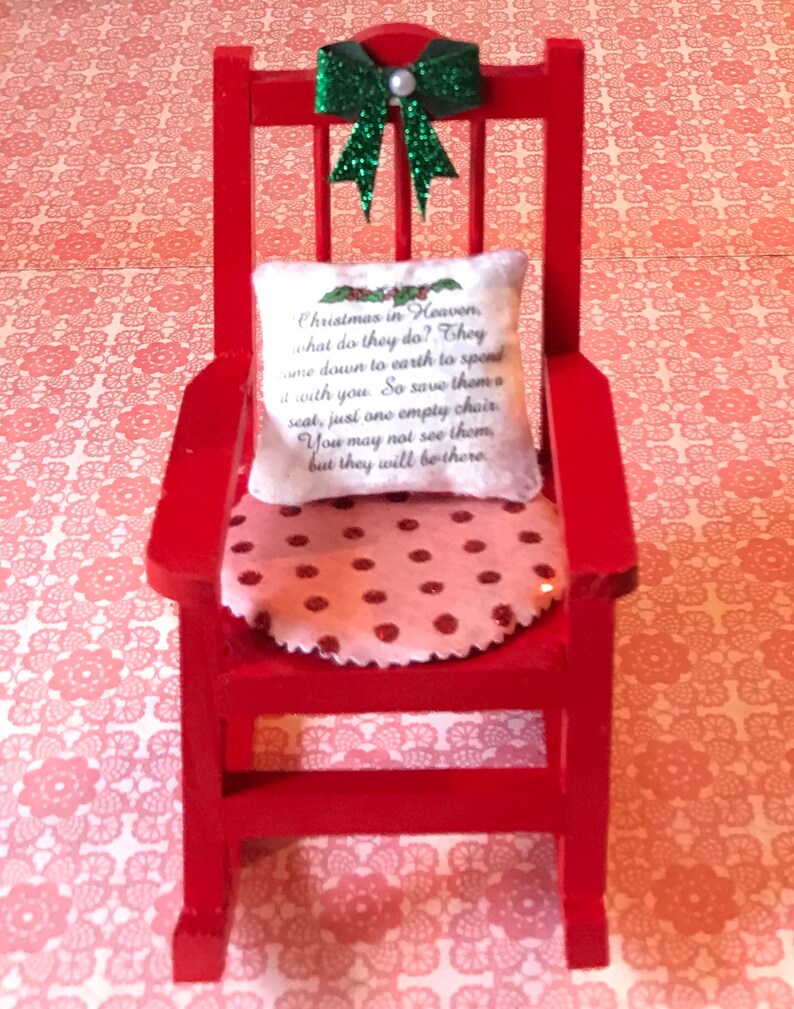 May include: A red rocking chair with a white cushion featuring the text "Christmas in Heaven, what do they do? They come down to earth to spend time with you. So save them a seat, just one empty chair. You may not see them, but they will be there." The chair has a pink and white polka dot cushion and a green glitter bow on the back.