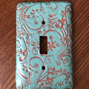 May include: A light blue ceramic light switch cover with a decorative copper floral pattern.
