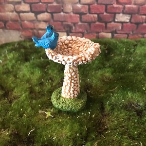 K&ouml;nnte beinhalten: Ein Miniatur-Vogelbad mit wei&szlig;er, strukturierter Oberfl&auml;che und gr&uuml;nem Sockel. Zwei blaue V&ouml;gel sitzen auf dem Rand des Bades.