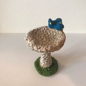 K&ouml;nnte beinhalten: Ein kleines wei&szlig;es Vogelbad mit strukturierter Oberfl&auml;che und gr&uuml;nem Sockel. Zwei blaue V&ouml;gel mit gelben Schn&auml;beln sitzen auf dem Rand des Bades.