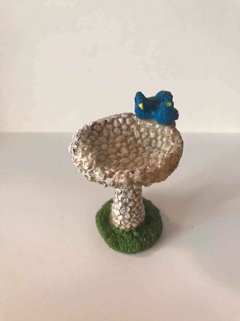 K&ouml;nnte beinhalten: Ein kleines wei&szlig;es Vogelbad mit strukturierter Oberfl&auml;che und gr&uuml;nem Sockel. Zwei blaue V&ouml;gel mit gelben Schn&auml;beln sitzen auf dem Rand des Bades.