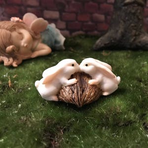 May include: Two white bunny figurines sharing a brown, textured, half-shell nut. The bunnies are facing each other and appear to be looking at the nut.