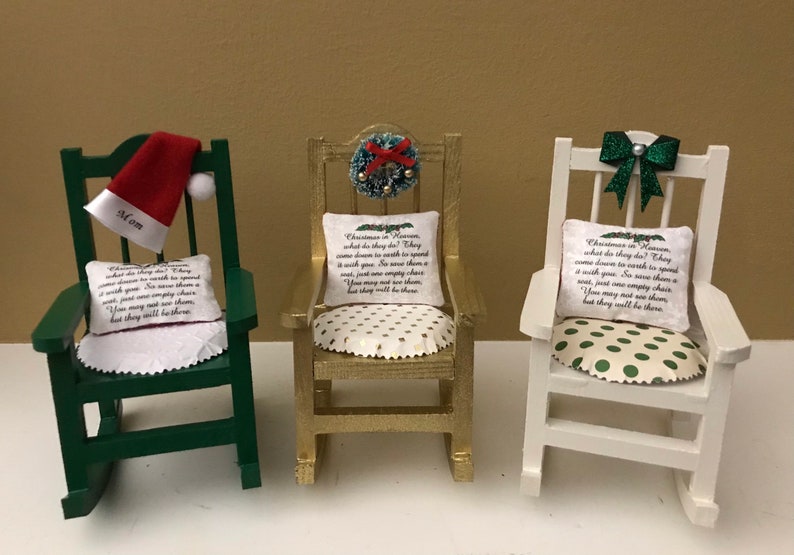 May include: Three miniature rocking chairs decorated for Christmas. The chairs are painted green, gold, and white. Each chair has a pillow with a Christmas poem printed on it. The green chair has a Santa hat on the back, the gold chair has a wreath with red berries, and the white chair has a green bow.