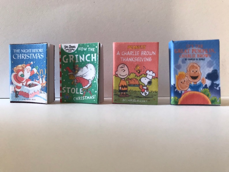 May include: Four miniature hardcover books with colorful covers. The books are titled "The Night Before Christmas", "How the Grinch Stole Christmas!" by Dr. Seuss, "A Charlie Brown Thanksgiving" by Charles M. Schulz, and "It's the Great Pumpkin, Charlie Brown" by Charles M. Schulz.