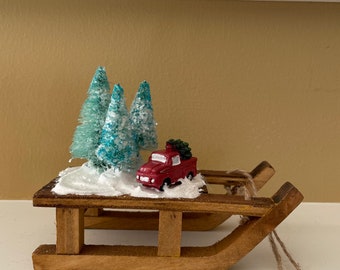 Puede incluir: Un adorno de trineo de madera con un camión rojo que lleva un pequeño árbol verde, rodeado de tres pequeños pinos azules y blancos escarchados. El trineo está decorado con nieve blanca y tiene un colgador de cuerda.
