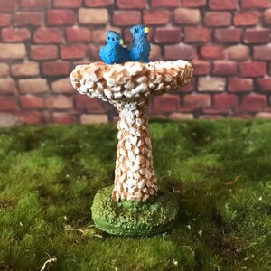 K&ouml;nnte beinhalten: Ein wei&szlig;er Miniatur-Vogelbad mit strukturierter Oberfl&auml;che, auf dem zwei blaue V&ouml;gel sitzen. Das Vogelbad steht auf einer gr&uuml;nen Grasfl&auml;che.