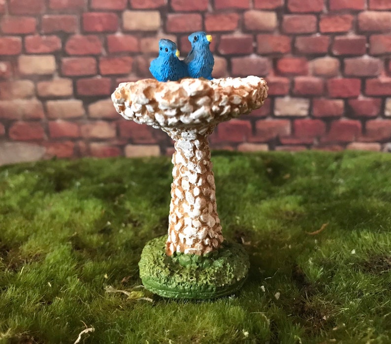 K&ouml;nnte beinhalten: Ein wei&szlig;er Miniatur-Vogelbad mit strukturierter Oberfl&auml;che, auf dem zwei blaue V&ouml;gel sitzen. Das Vogelbad steht auf einer gr&uuml;nen Grasfl&auml;che.