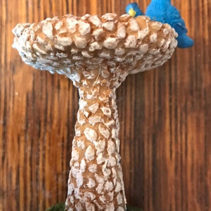 K&ouml;nnte beinhalten: Ein Miniatur-Vogelbad aus brauner und wei&szlig;er, gesprenkelter Keramik. Das Bad ist in Form eines Pilzes mit einer breiten, flachen Oberseite und einem hohen, schlanken Stiel. Zwei kleine blaue V&ouml;gel sitzen auf dem Rand des Bades.