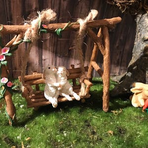 May include: Two white bunny figurines sitting on a wooden swing set with a green leafy vine with pink flowers. The swing set is made of natural wood branches. A third bunny figurine is sitting on the ground next to the swing set.