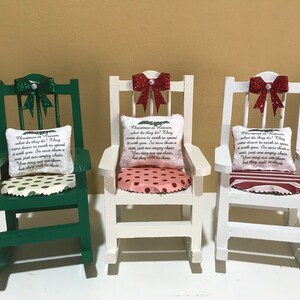 May include: Three miniature rocking chairs with red bows and pillows featuring a Christmas in Heaven theme. The chairs are painted green and white.
