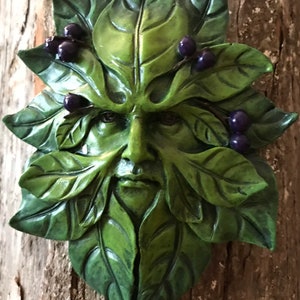 May include: Green Man wall sculpture with a face carved into leaves and berries. The sculpture is made of a green material and has a rustic look.