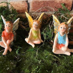 Op de afbeelding: Drie miniatuur fee-figuurtjes met vleugels, zittend op een bed van groen mos. De feeën dragen kleurrijke jurken en hebben geschilderde gezichten.