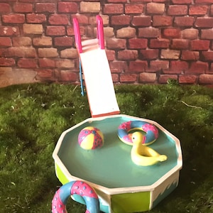 May include: A miniature plastic pool with a slide, a yellow duck, and two colorful inflatable rings. The pool is green and blue with a white rim. The slide is pink and white. The duck is yellow and has a blue beak. The rings are pink and blue with white polka dots.