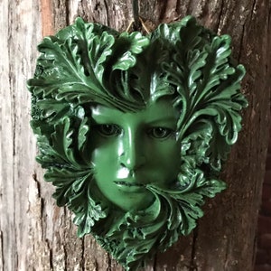 May include: A green, heart-shaped wall hanging with a woman's face surrounded by leaves. The face has a serious expression and is made of a textured material.