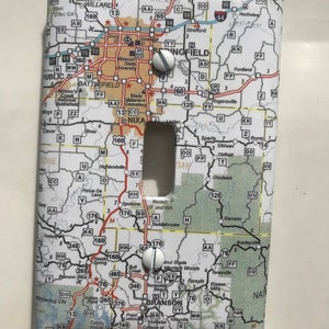 Pode incluir: Uma tampa de interruptor de luz com um mapa do Missouri com o texto "BRANSON" destacado. A tampa é branca com um mapa de cor marrom claro impresso nela. Há dois parafusos prateados nos cantos superiores da tampa.