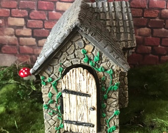 Fairy garden house with hinged door that opens is gray cobblestone with round windows, brick chimney and ivy framing the door