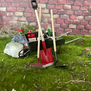 Puede incluir: Una pala roja en miniatura, un rastrillo y una azada se apoyan en una carretilla de madera en miniatura. La carretilla está llena de herramientas de jardinería en miniatura y una pequeña caja con la etiqueta "Tomate". Las herramientas están sobre un lecho de musgo verde.
