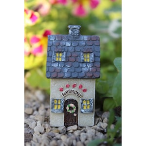 Op de afbeelding: Een miniatuur feeënhuis met een grijs dak van shingles, witte muren en een bruine deur. Het huis heeft twee ramen met gele ruiten en een bord met de tekst "Alleen voor feeën".
