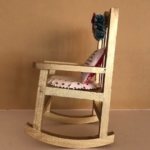 May include: A gold rocking chair with a white and red patterned cushion and a green wreath with red accents hanging on the back.