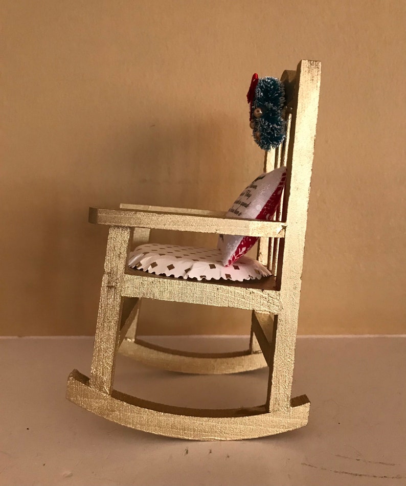 May include: A gold rocking chair with a white and red patterned cushion and a green wreath with red accents hanging on the back.