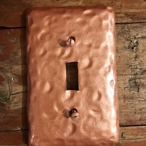 Puede incluir: Una tapa de interruptor de luz de color cobre con una superficie texturizada. La tapa tiene una sola abertura para el interruptor y dos agujeros para tornillos.