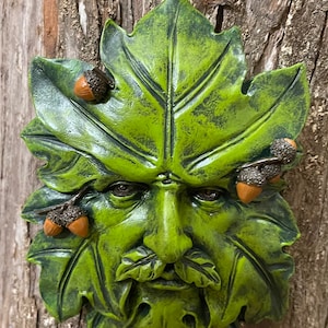 May include: Green man face sculpture with a leaf design and brown acorns. The sculpture is made of a textured material and has a rustic look.