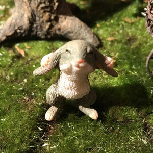 May include: A small gray and white rabbit figurine with closed eyes, sitting on a bed of green moss.