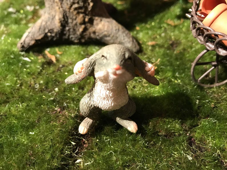 May include: A small gray and white rabbit figurine with closed eyes, sitting on a bed of green moss.
