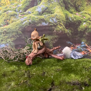 Op de afbeelding: Een klein, groenhuidig ​​feebeeldje met vleugels zit op een houten tak. De fee draagt ​​een bruine tuniek en heeft een droevige uitdrukking op zijn gezicht. Het beeldje is geplaatst tegen een achtergrond van met mos bedekte rotsen en bladeren.