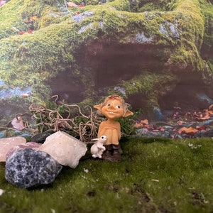 Könnte beinhalten: Eine kleine, lächelnde Gnomfigur mit spitzen Ohren und einem braunen Hut sitzt auf einem Bett aus grünem Moos. Der Gnom hält eine kleine weiße Hasenfigur. Der Hintergrund ist eine moosbewachsene grüne Waldszene.