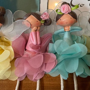 May include: Four fairy figurines with wings and floral accents. Each fairy is wearing a different color dress: yellow, pink, blue, and green. The fairies are sitting on a wooden surface.