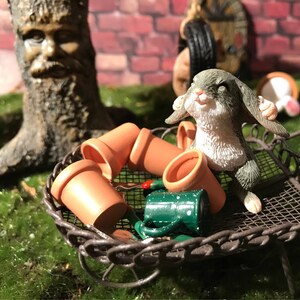 May include: A miniature gray rabbit figurine with white fur on its chest and belly, sitting in a brown wire basket with terracotta flower pots and a green watering can.