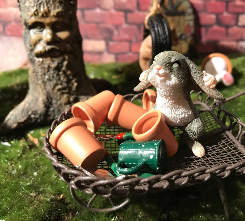 May include: A miniature gray rabbit figurine with white fur on its chest and belly, sitting in a brown wire basket with terracotta flower pots and a green watering can.