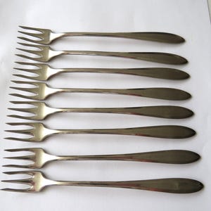 May include: A set of eight silver-toned metal forks arranged in a parallel vertical column. Each fork features a three-tined head and a smooth, tapered handle with a rounded end, reflecting a polished, metallic finish across the entire set.