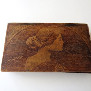 May include: A rectangular, antique wooden box with a carved profile of a woman. The woman's hair is styled in an updo, and the box has a decorative, arched border. The wood has a warm, brown tone.