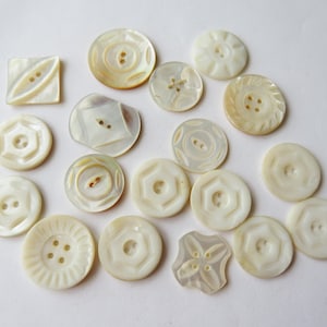 May include: A collection of off-white, iridescent buttons in various shapes and sizes. The buttons include round, square, and uniquely shaped designs, each with two or four holes for sewing. Some buttons feature embossed patterns, adding texture and detail to the collection.