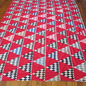 May include: A red quilt with a repeating pattern of triangles. Each triangle is bordered by red fabric and filled with a variety of patterned fabrics in black, white, blue, and tan. The quilt is laid out on a wooden floor.