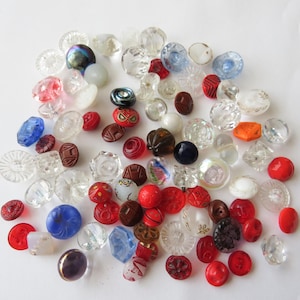 May include: A collection of assorted vintage buttons in various sizes, shapes, and colors. The buttons include clear, red, blue, and brown tones, with some featuring intricate designs and textures. A variety of materials are present, including glass and plastic.