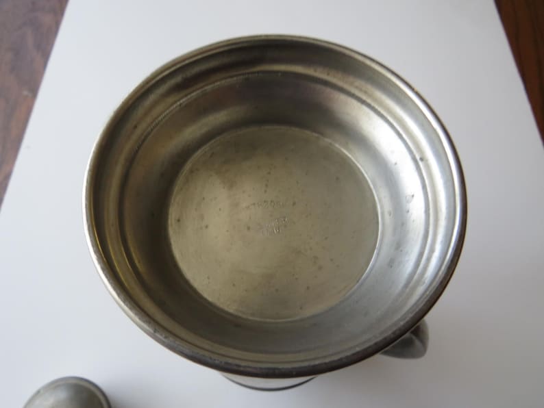 May include: A vintage pewter bowl with a handle. The bowl has a wide rim and a slightly textured surface. The interior is smooth and shows signs of age. The bowl is likely used for serving or display.