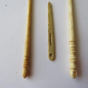 May include: Two long, slender, cream-colored carved bone tools with rounded, textured handles and a single flat, pointed bone needle with a central slit and a circular eye hole positioned between them, all featuring a natural, porous surface texture.