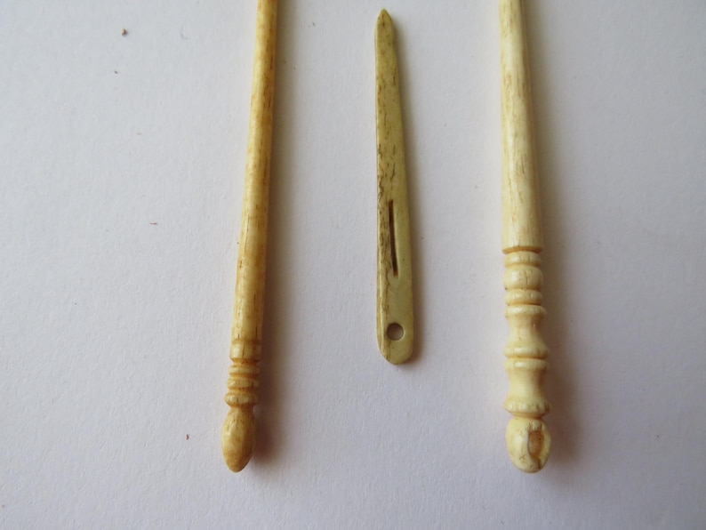 May include: Two long, slender, cream-colored carved bone tools with rounded, textured handles and a single flat, pointed bone needle with a central slit and a circular eye hole positioned between them, all featuring a natural, porous surface texture.