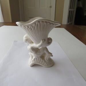 K&ouml;nnte beinhalten: Eine wei&szlig;e Keramikvase mit einer Cherubfigur, die ein muschelf&ouml;rmiges Gef&auml;&szlig; h&auml;lt. Die Vase hat ein strukturiertes, geripptes Design auf der Muschel und steht auf einem dekorativen Sockel. Der Gesamtstil ist klassisch und dekorativ.