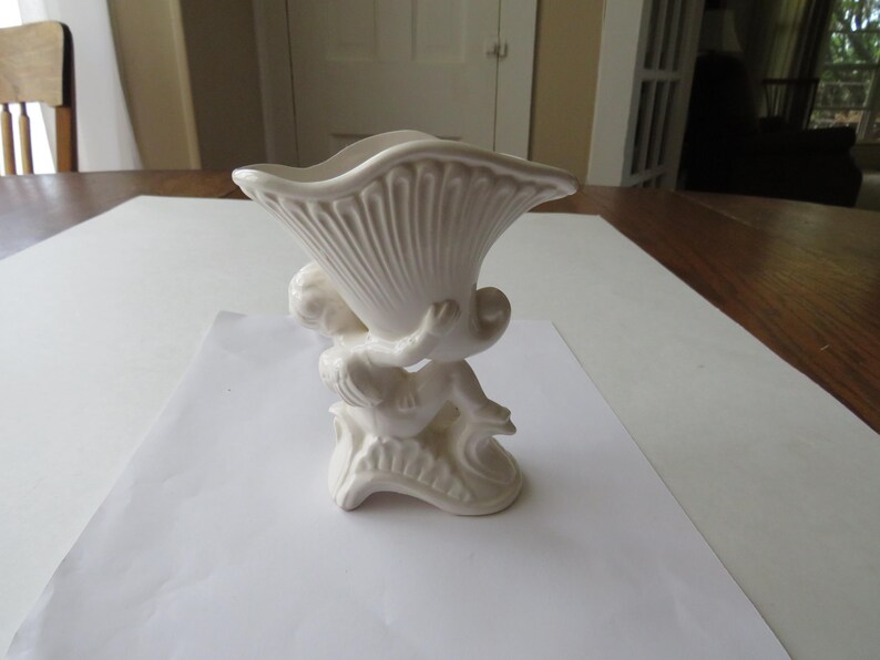 K&ouml;nnte beinhalten: Eine wei&szlig;e Keramikvase mit einer Cherubfigur, die ein muschelf&ouml;rmiges Gef&auml;&szlig; h&auml;lt. Die Vase hat ein strukturiertes, geripptes Design auf der Muschel und steht auf einem dekorativen Sockel. Der Gesamtstil ist klassisch und dekorativ.